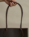 Brown leather tote bag held by a hand against a beige background
